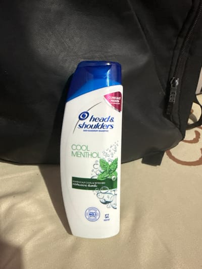 A white bottle of Head & Shoulders Cool Menthol Anti-Dandruff Shampoo is placed on a beige fabric surface in front of a black bag. The bottle has a blue cap and features branding and product information. A pink label on the bottle states "CLINICALLY PROVEN TECHNOLOGY." The shampoo is designed to leave the scalp cool and refreshed. A circular emblem on the lower part of the bottle reads "WORLD'S NO.1 SHAMPOO." The background suggests an indoor setting, possibly a room. There are no people visible in the image, and no specific landmarks identify the location as Manila, Philippines.