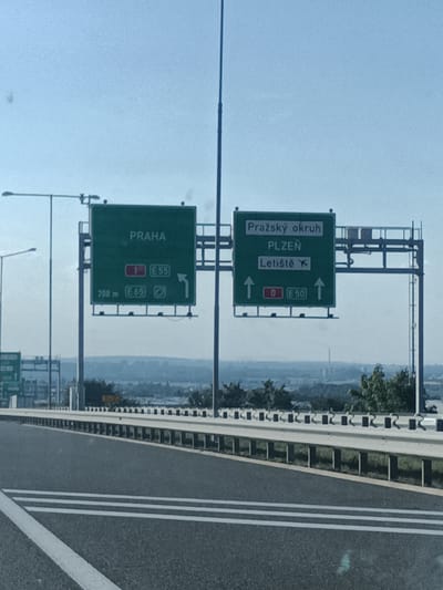The image shows a highway scene with overhead road signs in Czech Republic, likely near Kuří. The primary subjects are two large green directional signs.
The left sign indicates "PRAHA" (Prague) and shows a lane choice with "1 E 55" to the right. Below that, it specifies "200 m E 65" with an icon of a car.
The right sign points towards "Pražský okruh" (Prague Ring Road) and "PLZEŇ" (Pilsen). It also indicates "Letiště" (Airport) with an airplane icon. Below this, there are lane choices with "0 E 50" to the right, indicated by an arrow pointing straight up.
The scene is set on a clear day with a bright sky, suggesting daytime. The road appears to be a multi-lane highway with guardrails. In the background, a hazy landscape with trees and distant buildings can be seen, hinting at a semi-urban or suburban area. The overall impression is one of travel and navigation on a major road. No people are visible in the image.