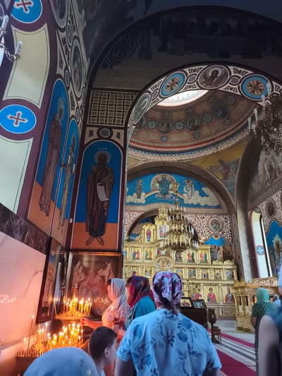 The photograph depicts the interior of the Church of the Ascension of the Lord in Ocnița, Moldova, captured from a low angle, looking towards the altar. The church features elaborate religious art, with frescoes adorning the walls and ceiling, depicting biblical scenes and figures. These artworks are characterized by vibrant colors, intricate details, and a predominantly blue and gold palette.
In the foreground, several individuals are visible, some in prayer or observing the religious service. They are dressed in everyday clothing, with women wearing headscarves. The lighting is primarily provided by numerous lit candles arranged in candelabra, creating a warm and intimate atmosphere. The altar area is dominated by a large, ornate iconostasis, a wall of icons separating the sanctuary from the nave, adorned with gilded elements and religious imagery. A large chandelier hangs from the ceiling, providing additional illumination. The floor is covered with a carpet featuring a striped pattern. The architectural style appears to be Eastern Orthodox, with arched ceilings and domes. The overall impression is one of reverence and spiritual devotion within a richly decorated religious space.