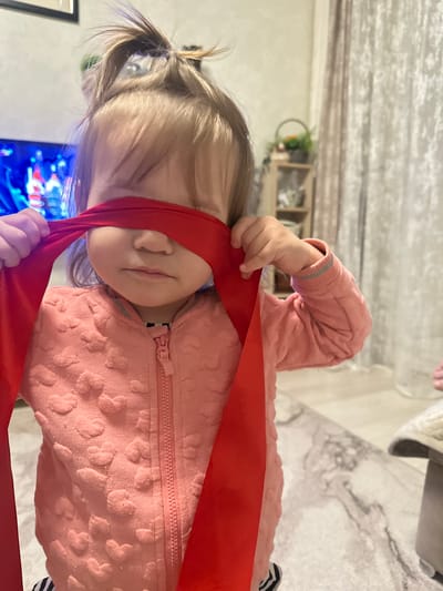 A young child stands indoors, blindfolded with a red cloth, wearing a pink heart-patterned sweater. The location is potentially in Uva, Russia. The background includes a patterned wall, shelves with plants, a television screen displaying blue light, and a light-colored floor. The child is holding the red cloth with both hands.