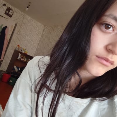 The image is a close-up, selfie-style shot of a young female with long, dark hair and brown eyes. She is wearing a white long-sleeved shirt. The background appears to be an interior setting, possibly a room. On the wall, there is a light fixture, a meter box, and some shelving units containing various items, including bottles, containers, and a pair of shoes. Two buckets, one red and one green, are visible on the floor in the lower left corner. The environment suggests a somewhat cluttered, lived-in space. The image does not provide any specific visual cues to determine the precise location within Baymak, Russia, other than implying it is an indoor residential or storage area.