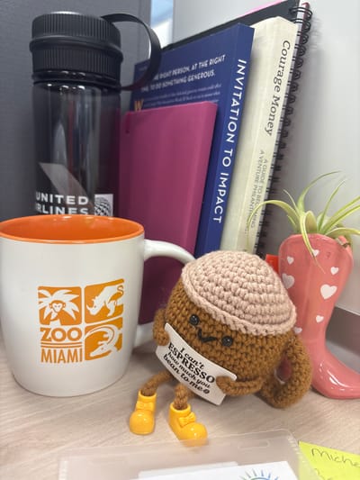 The image shows a desk scene with several objects, including books, a mug, a water bottle, and a small crocheted figure. The books are stacked, with the top one titled "Courage Money" and a subtitle reading "A Guide to Becoming a Venture Philanthropist." Below it is a blue book with the title "INVITATION TO IMPACT."

A white mug with an orange interior is prominent, decorated with orange graphic icons of animals, including a lion, an alligator, and a monkey, along with the words "ZOO MIAMI." A crocheted brown figure, possibly a bear or a squirrel, is perched on the edge of the mug. It wears a beige crocheted hat and a small white banner that reads "I can't ESPRESSO how much you bean to me." The figure has yellow crocheted boots.

A dark-colored water bottle with a black lid is also visible, lying horizontally. In the background, a pink, heart-decorated planter holds a small air plant. A yellow sticky note with handwritten text "Michele" is partially visible to the right. The overall scene suggests a workspace or a personal desk, with a casual and perhaps whimsical arrangement of items. The lighting indicates it is daytime. The visible text includes book titles, the mug's branding, and a humorous phrase on the crocheted figure. There are no people visible in the image.