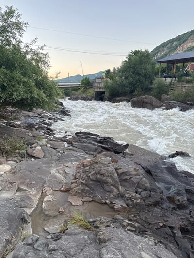 The image depicts a scenic view of a rushing river flowing through a rocky gorge in Kamennomostsky, Russia. The river, with its white, turbulent water, dominates the upper portion of the frame. The banks are lined with large, dark gray rocks, some appearing wet from the spray of the water, and interspersed with patches of green vegetation.
To the left of the river, a steep, tree-covered mountainside rises towards the sky. In the background, there are signs of human development, including buildings with dark roofs and a bridge crossing the river. Further in the distance, a street light and power lines are visible against the pale, hazy sky, suggesting either early morning or late evening. The sky itself is a soft, gradient hue, transitioning from a light blue at the top to a warm, peachy color near the horizon, indicative of sunrise or sunset.
The overall mood is one of natural beauty and raw power, with the dynamic flow of the river contrasting with the solid, ancient appearance of the rocks and mountains. There is no visible text in the image.