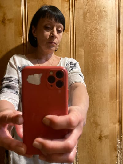 An adult female with short, dark hair and bangs is taking a selfie with a red smartphone. She is wearing a light-colored, patterned sweater and a thin necklace. She is standing in front of a wood-paneled wall. The image is cropped to focus on her face and upper body, and the smartphone is prominently featured in the foreground. The setting appears to be indoors, and the lighting suggests it may be evening. There is no indication that this scene is located in Yokohama, Japan.