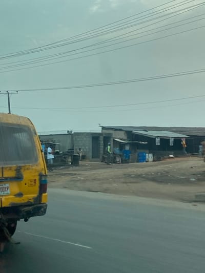 A roadside view shows a weathered yellow auto-rickshaw. Next to it is a row of modest structures, likely shops or workshops, with corrugated metal roofs and cinder block walls. Electrical wires crisscross the sky, adding to the utilitarian feel of the scene. The road is paved and appears slightly wet, contributing to the subdued light under an overcast sky. People are seen near the shops, engaged in activities that suggest a bustling yet commonplace street.