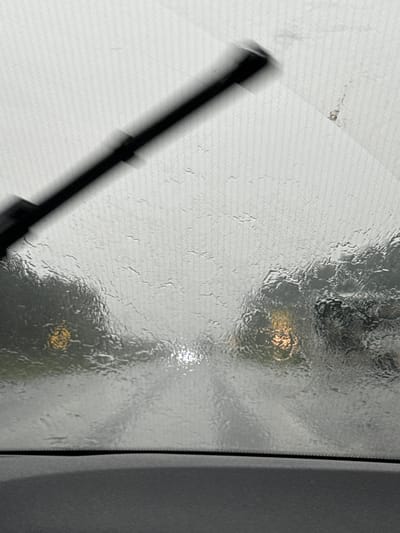 The image depicts a view from inside a vehicle looking through a rain-streaked windshield. A windshield wiper is visible in the upper left quadrant, blurred due to motion. The road ahead is visible, with headlights of oncoming vehicles creating bright, diffused spots through the water on the glass. The scene suggests inclement weather conditions, likely during daylight hours given the ambient light. The location is identified as Монетный, Russia.