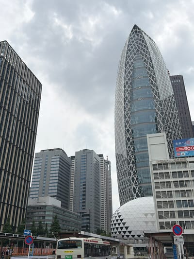 In Shinjuku, Japan, a wide-angle, low-angle shot captures a cityscape under an overcast sky. The foreground features a bus at a covered bus stop. To the left, a blue and white arrow sign points left, and a circular red and blue no parking sign is visible. A bus with a white exterior and red accents is stopped at the bus stop, displaying "ドライブレコーダー" and Japanese characters on its rear.

Dominating the mid-ground is a distinctive, shard-like skyscraper with a glass facade covered by a white lattice pattern. The words "MODE," "HAL," and "IKO" are visible vertically on the building's glass. To the left of this structure is a large, white geodesic dome.

In the background, several other skyscrapers of varying heights and architectural styles are present. One is a tall, dark rectangular building. Another, to the far left, is a slender, dark building with vertical white stripes. To the right of the geodesic dome, a white building with numerous visible staircases behind windows is present. Further to the right, a portion of a dark, rectangular skyscraper is visible, and a blue and white advertisement with Japanese text is displayed on a building. The overall scene depicts a modern urban environment.