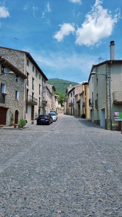 The image depicts a quiet, cobblestone street in the town of Montagut i Oix, Spain, under a bright, partly cloudy sky. Buildings with stone and stucco facades line both sides of the street, many featuring balconies and arched doorways. A dark blue car is parked on the left side of the street, followed by a white van. In the distance, a lush green hillside rises, indicating the town's rural or mountainous setting. The architecture suggests a historic European village. There are no people visible in the scene. The overall impression is one of tranquility and timelessness.