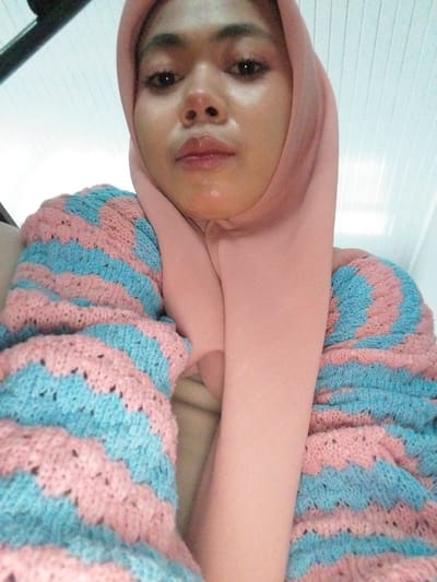 A close-up, low-angle, eye-level shot shows a person wearing a pink hijab and a knitted cardigan with alternating pink and blue chevron stripes. The person's face is partially visible, with their eyes looking directly at the camera. Their skin appears smooth with some shine, and their lips are slightly parted. The background consists of a white, ribbed ceiling or paneling. The image was taken indoors. No specific location in Tibojong, Indonesia is discernible from the visual information provided.