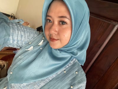 The image displays an indoor scene in Ketegan, Indonesia. The primary subject is a female, presented from the chest upwards, positioned slightly left of center, and looking directly at the camera. She is wearing a light blue satin hijab covering her head and neck, along with a matching light blue garment adorned with a textured or lace-like pattern, pleating, several white pearl beads, and two white butterfly appliqués on her left shoulder area. Her face exhibits applied makeup, including dark eyeliner, mascara, and brown lip color. In the background, to the left and lower, are the visible bare feet and lower legs of a second individual, who appears to be lying down on a patterned rug featuring red and brown tones. A light-colored handbag with a dark strap is also visible on the floor near the second individual. A dark brown wooden structure or panel occupies the right portion of the image.