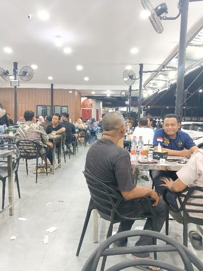 The image depicts a crowded, brightly lit casual dining establishment, likely a restaurant or food court, located in Seumadam, Indonesia, during nighttime hours. The environment features a grey tiled floor, a white ceiling with multiple recessed lights, and industrial-style black ceiling fans mounted on vertical poles. One side of the establishment appears enclosed with wood paneling, while the right side transitions into a semi-open area with a corrugated metal roof structure. Scattered debris, including paper, is visible on the floor.

Numerous individuals are seated at metal tables with black plastic chairs, engaged in eating and socializing. In the foreground, a man with short grey hair, wearing a dark grey t-shirt and dark pants, is seated with his back to the camera, his bare feet in flip-flops visible. To his right, another man with short dark hair, wearing a dark blue polo shirt, is seated at a table, smiling and looking directly towards the camera. Next to him, a third individual is partially visible. On their table are several bottled beverages (including what appears to be Aqua mineral water), a cup with a drink, and mobile phones.

In the midground and background, multiple groups of people, predominantly adults, occupy tables. One person on the left is wearing a patterned batik-style top. Other patrons are seen conversing, looking at their mobile phones, or consuming food and drinks. The overall scene indicates active social dining.