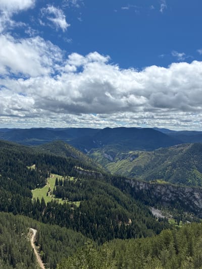 The image depicts a landscape with mountains, trees, and sky, located in Giovren, Bulgaria. The sky is partly cloudy with blue sky visible on the left side and a band of clouds down the center. Mountains are in the background, with foreground filled by forested hills. A small clearing with green grass is visible amidst the trees, with a small dirt road leading towards it.