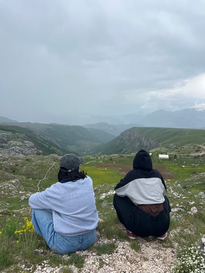 Two people are sitting at the edge of a mountainous area, overlooking a valley. The person on the left wears a grey hoodie and blue jeans, while the person on the right wears a black and grey hoodie and black pants. The landscape consists of grassy areas with rocks and yellow flowers, a valley, and mountains in the background. The location is Boričje, Montenegro. The weather appears overcast.