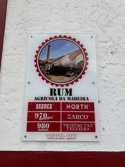 The image shows a sign for "Rum Agricola da Madeira" located in Porto da Cruz, Machico, Portugal. The sign is rectangular, mounted on a wall, and made of a clear material with white and red lettering. It features an image of a sugar cane mill. The sign lists different types of rum: "Branca," "970 Reserva," and "980 Madeira," and geographical names associated with rum production: "North," "Zarco," and "Tristão Vaz Teixeira." The sign also includes the name of the rum producer: "Engenhos do Norte." The wall behind the sign is painted white with a textured surface, while a red section is visible at the right edge.