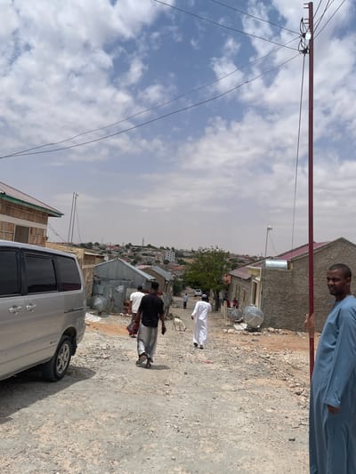 An eye-level, medium shot captures a dusty street scene in Hargeisa, Somaliland, under a partly cloudy sky. On the right, a man in a blue robe stands partially facing the camera, his expression unreadable. A red utility pole with wires and a lit bulb stands in the foreground. The street is unpaved and rocky, with a silver van parked on the left. Several people are walking down the street, including a man in a black shirt and patterned pants, another in a white robe, and a few others further in the distance. Goats wander near the people. Buildings with corrugated metal roofs and concrete walls line the street, suggesting a mix of residential and commercial structures. The background shows a dense collection of buildings stretching into the distance, characteristic of a city. The overall impression is one of everyday life unfolding in an arid urban environment.