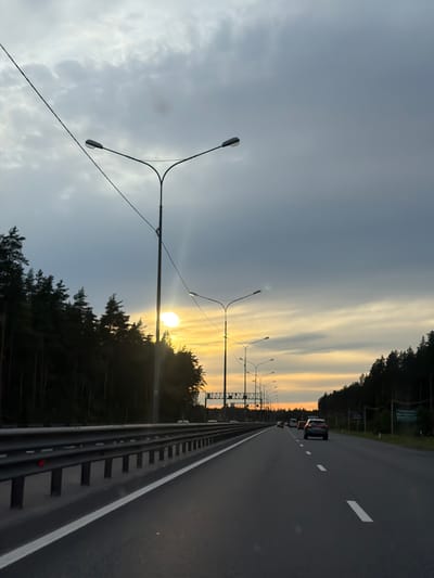 This media file depicts a road passing through a wooded area during sunset. The scene is shot from a moving vehicle, looking towards the right side of the road. The road itself is asphalt with white lane markings and a guardrail separating it from the shoulder. Several cars are visible on the road, driving in the same direction.
On the left side of the road, a series of streetlights with multiple arms line the edge of the highway. The sun is low in the sky, casting a warm, golden light that is creating dramatic streaks in the clouds. The sky is a mix of gray and yellow, indicating the transition from day to evening. The surrounding area is densely forested with tall pine trees.
The overall mood is serene and contemplative, with the natural beauty of the sunset contrasted with the man-made structure of the highway. The time of day is clearly sunset. There is no visible text in the image. The location is Okhtinskoye Razdol'ye, Russia.