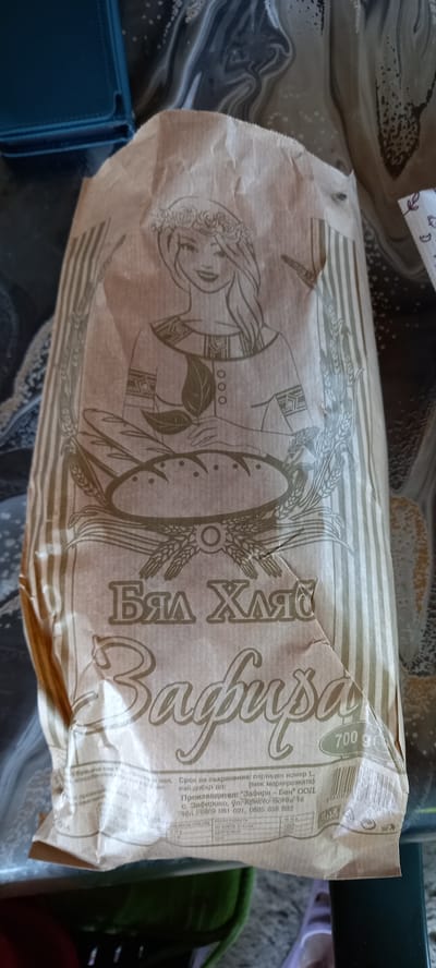 The image shows a close-up of a brown paper bread bag with Cyrillic text and a drawing of a woman wearing a flower crown and holding leaves. The woman is depicted sitting in front of a loaf of bread. The text on the bag reads "БЯЛ ХЛЯБ" (White Bread) and "Зафира" (Zafira), which is also likely the name of the bakery or brand. Below this, there is more text providing production information, including the manufacturer "ЗАФИРА - БЕН" ООД. The location "гр. Зафирово, ул. Христо Ботев 16" (Zafirovo village, Hristo Botev street 16) is visible. A barcode and nutritional information are also present on the lower part of the bag. The bag appears to be sitting on a patterned surface. No people are directly visible in the image, but the drawing implies a person. The context of the text indicates this is a product from Iskra, Bulgaria, specifically the village of Zafirovo.