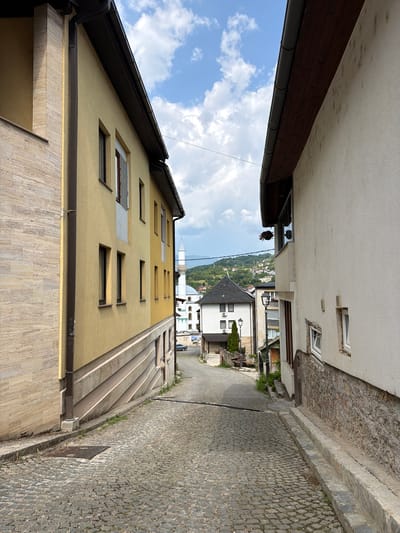 This is a rotated, eye-level, medium shot of a narrow cobblestone street in Jajce, Bosnia and Herzegovina. The street is lined with buildings, predominantly featuring light-colored stucco facades.
On the left, a yellow building with a dark-tiled roof dominates the foreground, displaying several small, rectangular windows. Above it, a beige building with a dark overhang and visible gutter system is partially visible. Further down the street on the left, more buildings with white and light-colored facades can be seen.
On the right side of the street, a stone wall runs along the edge of the cobblestone pathway. Several buildings of varying heights and architectural styles line this side, including a white building with a black-tiled roof and an air conditioning unit visible. A lamppost is also present on this side. In the distance, a white minaret and a dome are visible, suggesting the presence of a mosque. Greenery, in the form of trees and small plants, is interspersed between the buildings and along the street.
The sky is visible above the buildings, featuring blue skies with scattered white clouds. The lighting suggests it is daytime, likely mid-morning or afternoon, with bright sunlight casting shadows. The scene appears to be a quiet, residential or commercial area within a town. No people are clearly visible in the image, and no text is discernible. The overall mood is peaceful and somewhat historic, characteristic of European towns.