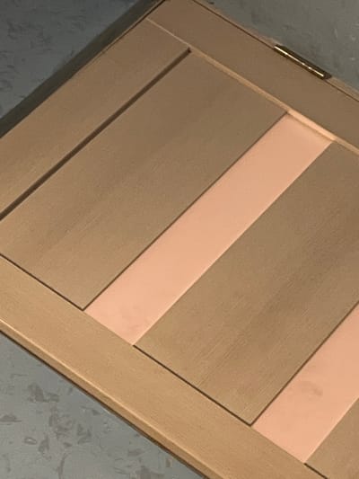 The image displays a close-up, angled view of a wooden surface with a diagonal design. The surface is divided into several panels, with alternating broad sections of light brown wood grain and narrower, lighter pink panels. A brass hinge is visible along the top edge of the wooden surface. The background is a dark gray textured surface. The scene appears to be indoors, likely a piece of furniture or cabinetry. There is no information within the image to suggest a location in Jrarbi, Armenia.