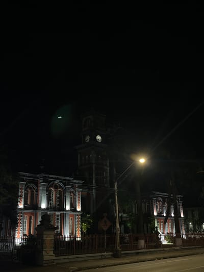 The image shows a building at night in Port of Spain, Trinidad and Tobago. The building has red and white trim and appears to have multiple floors. A street light is visible above the building, and there is a clock tower. The image is oriented sideways, with the building appearing on the right side of the frame. A fence is visible in the foreground. The night sky is dark and occupies the majority of the image. Based on the architecture and location, the building appears to be the Red House (Parliament Building).