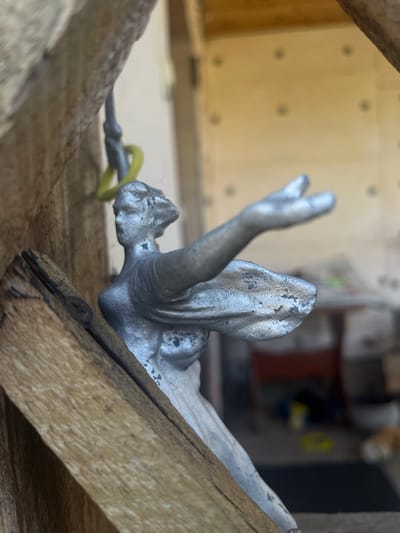 A weathered silver statuette of a winged female figure is positioned on a rustic wooden structure. The figure is seen in profile, with its head turned to the left and its right arm extended forward. A yellow cord is attached to the statuette, extending upwards and out of frame. The wooden structure appears to be part of an outdoor setting, possibly a shed or a fence, with rough, unfinished planks. In the background, the scene is blurred, suggesting an indoor or semi-outdoor environment with faint details of furniture and possibly a doorway or window. The overall impression is one of an old, possibly discarded or repurposed decorative item in a somewhat neglected or work-in-progress environment. The location in Preobragenskaya, Russia is not discernible from the visual information provided.