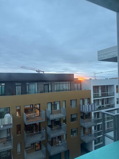 This photograph captures a residential building with balconies, located in Reykjavik, Iceland. The sun is setting, casting a warm orange glow across the sky and illuminating the windows of the building. Two cranes are visible in the distance, suggesting ongoing construction in the area. The building appears to be modern, with a mix of dark and light-colored facades and numerous balconies. The sky is overcast, with hints of blue peeking through the clouds. The overall scene depicts an urban landscape at dusk, with a focus on the architectural details of the building and the atmospheric quality of the sunset.