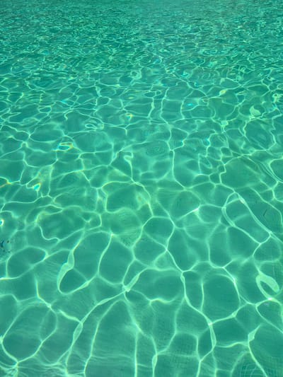 The image shows the sunlit surface of a swimming pool in Les Meravelles, Spain. The water is a clear, turquoise color, and the surface is disturbed by ripples, creating intricate patterns of light and shadow. The bottom of the pool is tiled, with a subtle grid pattern visible through the water. There are no people or objects in the water, and the focus is entirely on the texture and play of light on the water's surface. The overall impression is one of tranquility and the refreshing clarity of clean water.