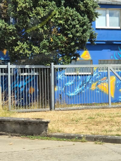 A blue building with a mural of abstract yellow and blue shapes and designs is visible behind a metal fence. A large green tree partially obscures the building and mural. The fence is made of vertical metal bars with a horizontal top railing. The ground in front of the fence is covered in dry, yellow grass, and a concrete curb and sidewalk are in the foreground. The building has at least two windows with horizontal blinds. The mural appears to feature fluid, swirling patterns in blue and yellow, with some geometric elements. The scene suggests an outdoor urban or suburban environment.