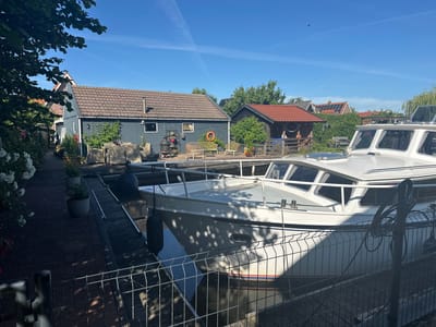 The image depicts a serene canal scene in Ossenzijl, Netherlands, under a clear blue sky. In the foreground, a white motorboat is docked alongside a wooden pier. The boat has a white hull with a blue stripe and a red trim line. Two dark fenders are attached to the boat's side, resting against the pier. A metal fence with a wire mesh pattern separates the viewer from the pier and the boat.
Behind the boat and pier, several buildings are visible. The largest is a dark blue structure with a brown tiled roof and a chimney. Adjacent to it is a smaller building with a red tiled roof. Both buildings appear to be houses or sheds, with windows and a chimney. Greenery, including trees and bushes, surrounds the buildings and lines the canal.
To the left of the buildings, a pathway made of brick pavers leads away from the canal. This pathway is lined with potted plants and a large bush with white flowers. The environment suggests a peaceful residential area, possibly a holiday park or a village along the water. The sunlight casts dappled shadows from the trees onto the boat and the pier, indicating it is daytime, likely mid-morning or afternoon. No people are visible in the image.