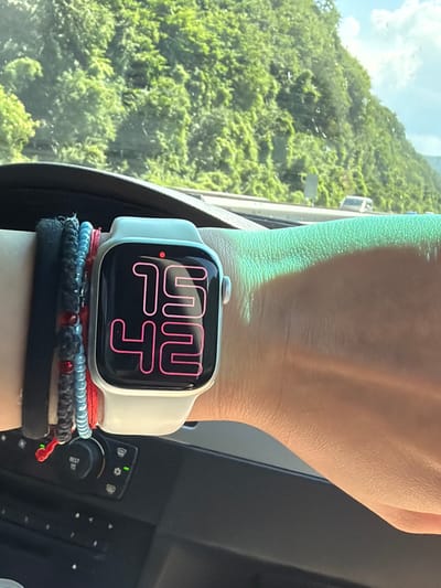 The image shows a close-up of a person's wrist wearing a white Apple Watch. The watch face displays a stylized digital time in pink font, reading "12:04". The watch is adorned with several bracelets underneath, including a red string bracelet, a blue beaded bracelet, and a dark grey beaded bracelet. The person's hand and wrist are positioned in front of a window, through which a lush green hillside covered in trees is visible under a bright, partly cloudy sky. The reflection in the window suggests the person is likely inside a vehicle, possibly a car or bus, given the presence of what appears to be dashboard controls to the right of the watch. The sunlight casts a bright glow on the person's skin and the watch. The overall impression is a snapshot of a moment during a journey, with the natural scenery outside contrasted with the modern technology on the wrist. No visible text is present other than on the watch face itself.
