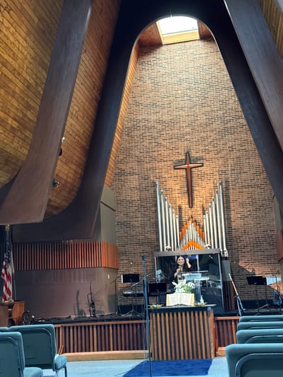 A view from the interior of a church, with an eye-level view of the wooden slatted floor, a curved wooden beam going towards the ceiling, and a brick wall with a cross and organ pipes above the stage. The pastor is at the podium.
The church appears to be located in Columbus, Ohio, USA.