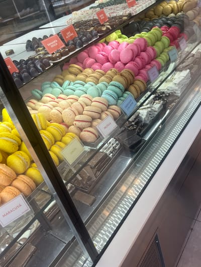 The image displays a brightly lit, multi-tiered glass and stainless-steel confectionery display case inside a commercial establishment in Tolhuin, Argentina. The case showcases various desserts and chocolates, meticulously arranged.
The upper shelves contain assortments of chocolates, including dark, milk, and white varieties. Specific sections are identified by small labels in Spanish: "BANANITA DULCE" (Sweet Banana), "CAFÉ AL COÑAC" (Coffee with Cognac), and "FRUTOS SECOS" (Dried Fruits).
Below the chocolates, multiple rows of macarons are precisely stacked by color and flavor. Visible flavors include hot pink, light green labeled "PISTACHO" (Pistachio), pink labeled "FRUTOS ROJOS" (Red Fruits), light brown, light blue, and cream-colored macarons with a red filling labeled "FRUTILLA" (Strawberry). The lowest visible tiers feature yellow macarons labeled "LIMÓN" (Lemon) and peach-toned macarons labeled "MARACUYÁ" (Passion Fruit).
The lower sections of the display case hold various chocolate and nougat products, including slabs of white chocolate, some labeled "CHOCOLATE CON LECHE" (Milk Chocolate), and dark chocolate bars, some with visible inclusions.
The environment appears clean and organized, with strong artificial lighting illuminating the products. Reflections are visible on the glass of the display case. No individuals are present within the visible frame, and no actions or interactions are occurring.