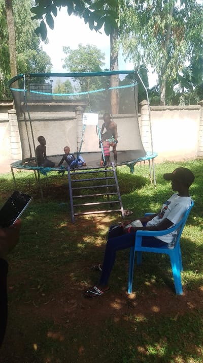The scene depicts an outdoor recreational area, presumably within Siaya Township ward, Kenya, on a sunny day. A large, black-matted trampoline with a blue-rimmed safety net stands centrally. A four-rung metal ladder leans against the trampoline, providing access.
Three children are present on the trampoline. One older child, shirtless and wearing colorful striped shorts (red, pink, and black), stands upright near the center, holding one of the trampoline poles. To the left, a younger, shirtless child in dark shorts sits on the trampoline surface. Further left, a very young child, dressed in a dark long-sleeved shirt with a white graphic and light shorts, reclines on the trampoline mat. A white rectangular sign with black text, largely illegible, is attached to the trampoline netting.
In the foreground, to the right, a person wearing a black baseball cap, a white t-shirt with a design, dark blue pants, and beaded sandals sits in a blue plastic chair, observing the trampoline. This individual has a pink bracelet on their right wrist. To the immediate left in the foreground, a hand holds a black smartphone with a visibly cracked screen.
The ground beneath and around the trampoline is a mix of reddish-brown soil and green grass, with clear indications of sunlight and shadow. A light-colored concrete wall or fence with pillars is visible in the mid-ground, behind the trampoline. The background consists of dense green foliage and tall trees under a bright sky. Overhead leaves from an unseen tree are visible at the top center of the frame.