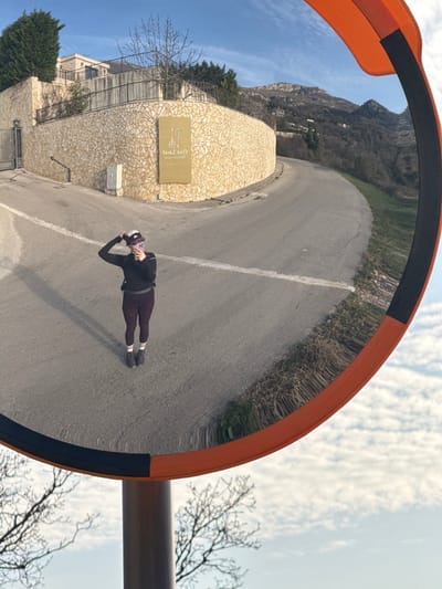 A convex traffic mirror with an orange and black frame reflects a outdoor scene. The central subject is a female individual standing on a paved asphalt road, directly facing the mirror. She is wearing a dark long-sleeved top, dark leggings, dark shoes, and white socks with two horizontal stripes. A small dark backpack is visible on her back. Her right hand holds a mobile phone, and her left hand is raised to her head, adjusting a dark baseball cap/visor. Her elongated shadow is cast to her left on the road surface.
Behind the individual, the paved road curves sharply to the right and ascends. A single white line is painted horizontally across the road. To the left of the road, a substantial retaining wall constructed from light-colored, irregularly shaped stones supports a building that is partially visible behind it. A metal gate is present on the far left. Affixed to the stone wall is a rectangular, light-brown sign with inverted text, clearly legible as "Oak Leaf" with stylized tree/leaf designs, and "Restaurant" below it. This establishment is located in Vojnići, Montenegro. Beyond the curving road and structures, distant mountains covered with sparse vegetation rise under a clear blue sky punctuated by scattered white clouds. The lower portion of the mirror reflection reveals a segment of a tree with bare branches against the sky. The scene is illuminated by direct sunlight, indicated by the distinct shadow of the person.
