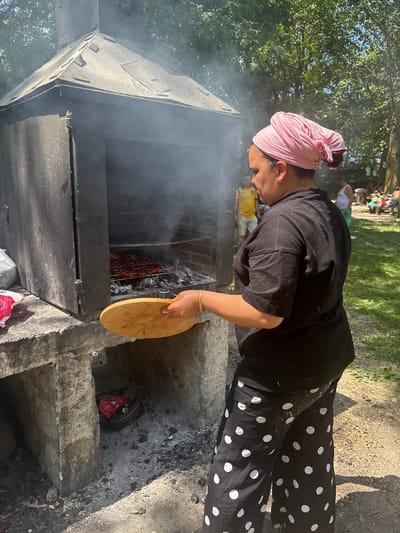 A woman, wearing a pink headscarf and a black t-shirt with white polka dot pants, stands in front of a large, dark-colored barbecue grill. She is holding a round wooden board, and smoke billows from the grill. Inside the grill, racks of food, appearing to be sausages or meat, are cooking over embers. The barbecue is made of concrete and has an opening at the bottom. The scene appears to be outdoors, with trees and a grassy area visible in the background. Other people are present in the background, suggesting a gathering or event. The location is identified as Campezo / Kanpezu, Spain.
