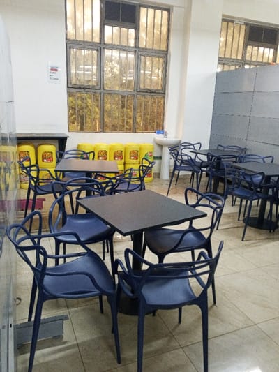 The photo depicts a brightly lit interior with several tables and chairs. Yellow containers are stacked near a window with a view of trees. A sink is positioned near the window. A large, gray panel dominates the right side of the room. The floor is tiled. The chairs are blue and have an unusual design. The tables are dark and square.