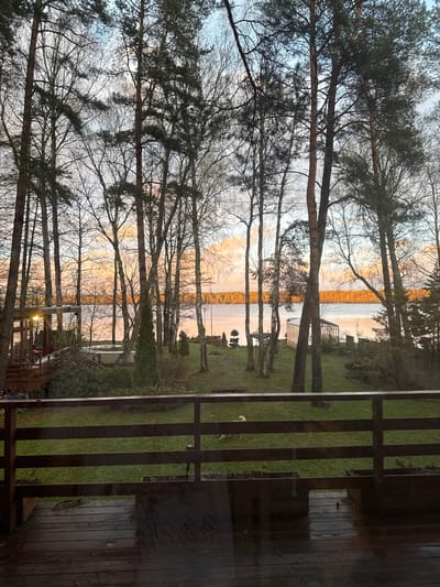 The image shows a view from a residential property in Baltezers, Latvia, towards a lake. Tall trees line the shore, and their branches obscure the sunset sky. The lake reflects the colors of the sunset. A wooden deck and fence are visible in the foreground. No people are visible.