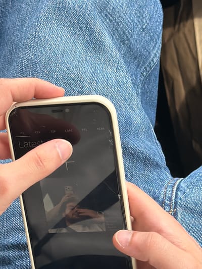 A person is holding a cracked screen smartphone over their blue denim-clad lap. The phone screen reflects the image of a person taking a photo. The location is not discernible from the given image.