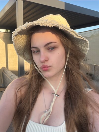 A young female with long brown hair and blue-green eyes is captured in a close-up selfie outdoors, wearing a straw bucket hat and a white ribbed tank top. She is puckering her lips for the camera and has a silver necklace with a cross pendant. The background shows a patio area with a modern pergola and lounge furniture, bathed in the warm, golden light of late afternoon or early morning. The lighting suggests it could be a sunny day in Slobozhanske, Ukraine, possibly on a residential patio or a public outdoor space.