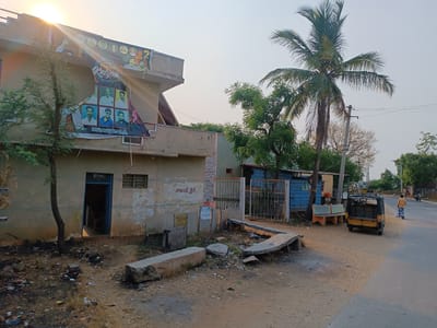 The image shows a street in Kothacheruvu, India, with a tan building on the left adorned with a large birthday banner for "PAWAN Kalyan" featuring multiple photographs. A tree partially obscures the banner. The building has an open blue doorway and a window. Debris litters the ground in front of the building. A metal gate stands between the building and a small, dilapidated structure with blue siding. A three-wheeled auto-rickshaw sits parked on the right side of a dusty road. A man walks along the right side of the road, wearing a yellow shirt and a patterned sarong-style lower garment. A tall palm tree is visible behind the blue structure. The sky is a hazy grey.