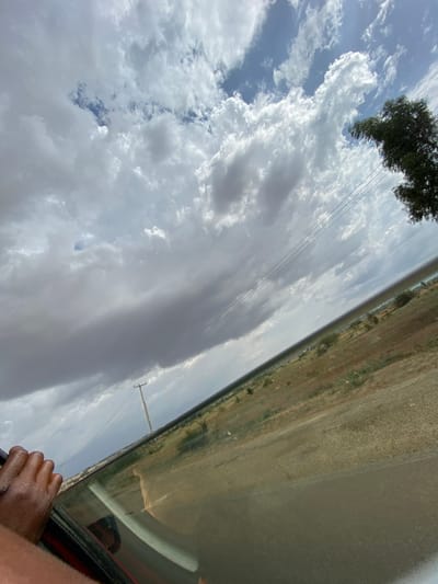 The image shows a view from inside a car, possibly moving. The foreground features a portion of the car window, frame, and a person's hand resting on the door. Reflected in the window is a person's face. The background is divided between the sky and landscape. The sky occupies a significant portion of the frame, filled with numerous white clouds, some areas showing patches of blue sky. A tree is seen in the upper left. Power lines traverse the sky. The landscape is a flat expanse of dry, grassy terrain. Based on the location provided (Barakin Columbai, Nigeria), the environment likely reflects a semi-arid or savannah climate. There is no direct evidence of specific subjects, objects, actions, or interactions aside from the elements described above. The photograph appears to capture the scene while traveling on a road.