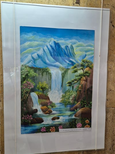 A framed painting of a mountain landscape with waterfalls hangs indoors. The painting features stylized blue and white mountains on the left, flowing into waterfalls cascading through rocky cliffs with lush vegetation and flowers on the right. A small label is affixed to the right side of the painting, containing text in a language that is not discernible in the provided image. The frame is white, and a white string hangs across the top of the frame. The background appears to be a textured surface. The location of Srebarna, Bulgaria, is not apparent.