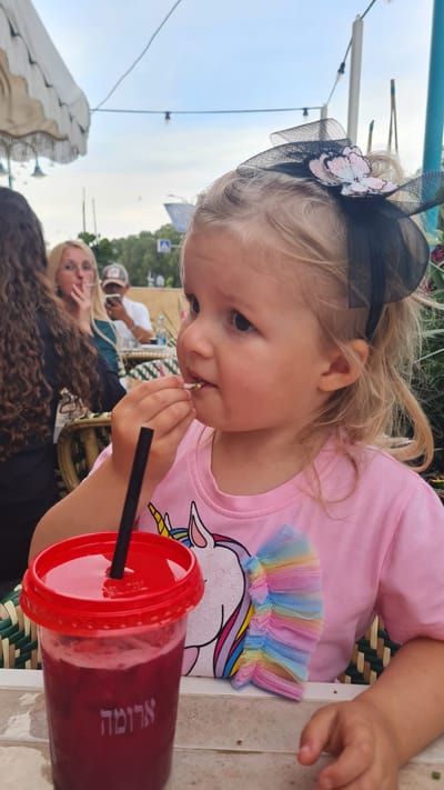 A young child sits at a table outdoors in Kiryat Gat, Israel. She wears a pink shirt with a unicorn design and a black bow headband with a flower on it. She is eating something from her hand. A large red cup with a straw stands on the table. In the background, other people are visible, including a woman with long curly hair and a man sitting at another table. The sky is visible in the background. The name 'Aroma' is written on the red cup.