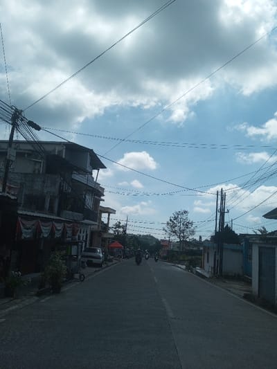The image displays a daytime street scene in Naung Bo Oe, Myanmar. A paved asphalt road, featuring a faded white broken center line, extends into the distance under a partly cloudy blue sky. Numerous black utility lines are visible crisscrossing overhead.

On the left side of the road, a multi-story concrete building with visible wear dominates the foreground. Red and white banners are affixed to its lower level. A small, wheeled cart and potted plants are situated on the sidewalk area. A light-colored sedan is parked further down the road on the left. Beyond the parked sedan, a red canopy tent is set up in front of an additional structure. A utility pole with a dense cluster of wires stands prominently on the far left.

On the right side of the road, another multi-wire utility pole is visible. Further along, a low, light-colored building or wall with a blue door or gate is present.

Several individuals are operating motorcycles, traveling away from the viewer down the road. In the background, hills covered with vegetation are visible under the sky.
