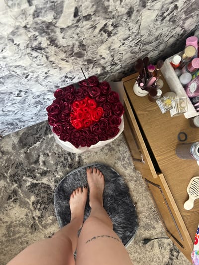 The image depicts an indoor scene, likely a bedroom or vanity area. A bouquet of red and burgundy roses is prominently displayed, arranged in a heart shape in the center of the bouquet. To the right of the roses, a wooden dresser or vanity holds various cosmetic items, including makeup brushes in a white holder, perfume bottles, and other small containers. A white fluffy towel is draped over the edge of the dresser. On the floor, a person's feet are visible, standing on a gray, furry mat. The feet have painted toenails. A tattoo is visible on the person's thigh. The background features a wall with a marbled pattern. The lighting suggests an indoor setting, possibly with natural light from a nearby window. The location is stated to be Blizhnii Khutor, Moldova, though there are no specific visual cues in the image to confirm this.