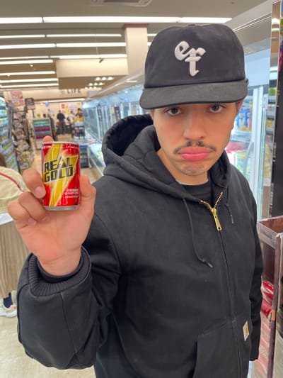 A man stands in a brightly lit convenience store in Nakano, Japan, holding a can of "Real Gold" energy drink. He wears a black jacket and a black cap with white lettering. Shelves of products are visible in the background. The store, labeled "LIFE," has bright fluorescent lighting and rows of consumer goods.