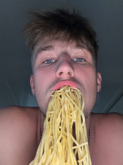 This is a close-up, head-and-shoulders, eye-level shot of a young, light-skinned man with short, light brown hair and blue eyes. He is holding a large amount of cooked spaghetti in his mouth, with the noodles spilling out. He is shirtless, and a silver chain necklace is visible on his chest. The background is dark and appears to be a plain wall or curtain. The image is poorly lit, and the focus is on the man's face and the spaghetti. There is no indication of the location in the city of Vanino, Russia, within the image itself.