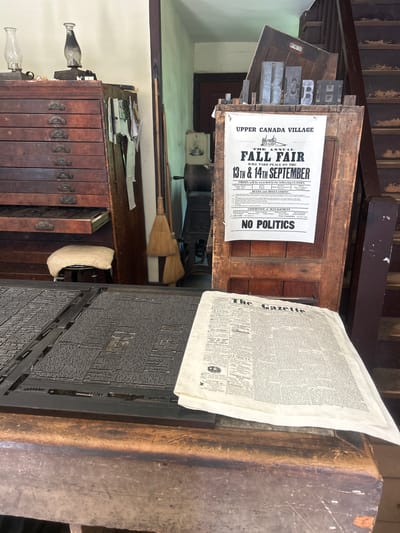 This image depicts a historical printing press setting within Upper Canada Village, South Dundas, Canada. The scene shows a wooden cabinet with several drawers, possibly used for storing printing materials. On top of the cabinet, a large piece of letterpress printing equipment is visible, covered with small metal characters used for setting type.

Prominently displayed is a poster advertising the "Annual Fall Fair" of Upper Canada Village, scheduled for the 13th and 14th of September. The poster also lists prize categories, such as "Agricultural Products" and "Dairy Products," and the "Committee of Management" and "Secretary." A large, folded newspaper, identified as "The Gazette," is also present, leaning against the printing equipment.

In the background, a wooden door or partition is visible, holding a collection of large metal letters, likely part of a larger printing setup. A broom made of straw leans against the wall, suggesting a rustic or well-used workspace. The overall impression is of a working historical exhibit, showcasing the tools and activities of a bygone printing era. The lighting suggests it could be daytime, with the scene illuminated by natural or artificial light sources within the building. There are no people visible in the image.