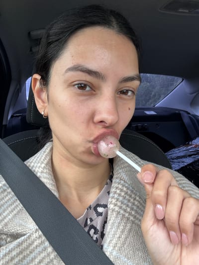 A woman with dark hair and light brown eyes is in the driver's seat of a car, wearing a beige and gray plaid jacket over a leopard-print top. She is holding a lollipop with red and green specks to her lips. A gray seatbelt is visible across her chest. The background shows the car's interior and a dark, out-of-focus exterior, suggesting an overcast day. The location is not specified in the image.