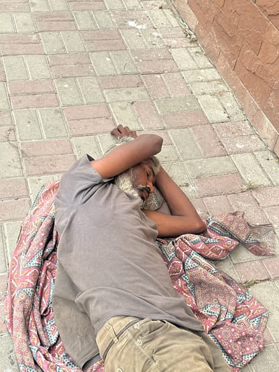 An elderly man sleeps on a patterned blanket on a sidewalk composed of rectangular paving stones, adjacent to a red brick wall. The man is lying on his back with his arms covering his face. He is wearing a grey t-shirt and khaki pants. The image appears to have been taken outdoors in a city.